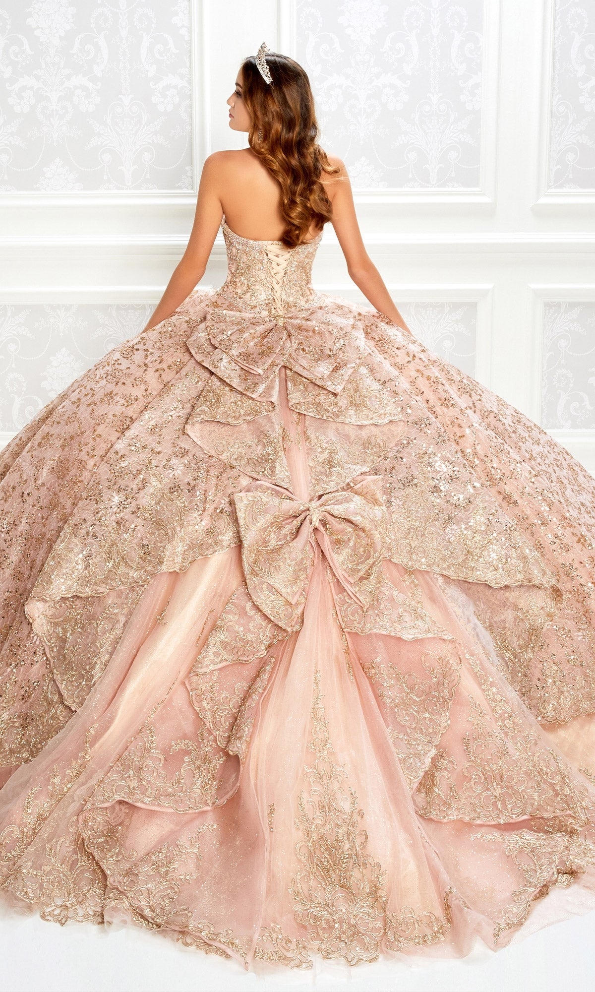 A woman in the Strapless Quinceanera Dress: Princesa PR22022—blush pink with gold details, large bows, and a tiara—stands facing away in an ornate room.